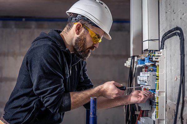 A male electrician works in a switchboard with an electrical connecting cable, connects the equipment with tools.